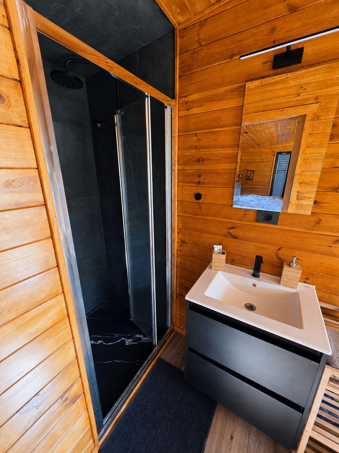 Walk-in shower cabin next to the bathroom vanity, finished in dark tile and timber