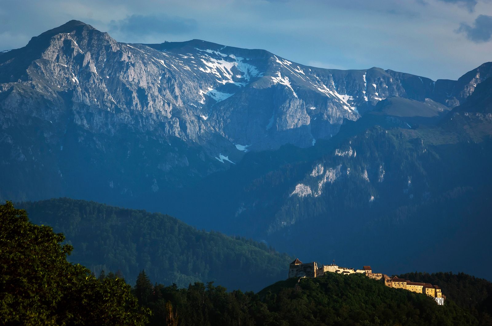 Râșnov Citadel lit at sunset on its hilltop, with the snow-streaked Bucegi mountains behind and the hillside RÂȘNOV sign below the walls